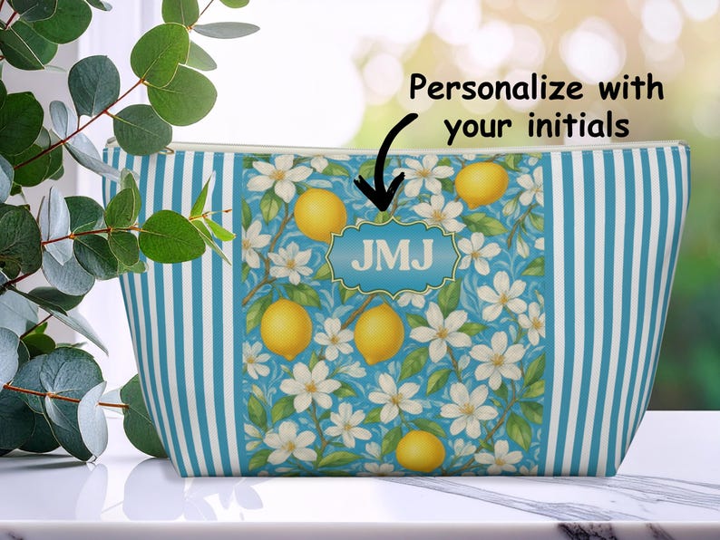 May include: A zippered cosmetic bag with a blue and white striped pattern. The center panel features a floral design with lemons and white flowers. The bag has the initials "JMJ" in a blue, decorative frame. The text "Personalize with your initials" is above the bag.