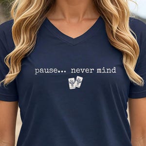 Pode incluir: Uma t-shirt azul-marinho com decote em V, apresentando o texto branco "pause... never mind" estampado no peito, acompanhado por um pequeno gr&aacute;fico de duas canecas brancas de estilo tiki posicionadas abaixo do texto no centro da t-shirt.