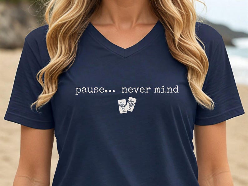 Pode incluir: Uma t-shirt azul-marinho com decote em V, apresentando o texto branco "pause... never mind" estampado no peito, acompanhado por um pequeno gr&aacute;fico de duas canecas brancas de estilo tiki posicionadas abaixo do texto no centro da t-shirt.