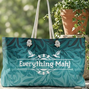 May include: A teal tote bag with a floral design and white rope handles. The bag has the text "Everything Mahj" in white, with two bird illustrations. The bag is designed for carrying items, suitable for shopping or leisure.