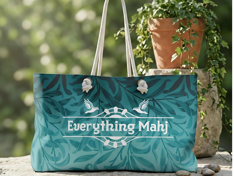 May include: A teal tote bag with a floral design and white rope handles. The bag has the text "Everything Mahj" in white, with two bird illustrations. The bag is designed for carrying items, suitable for shopping or leisure.
