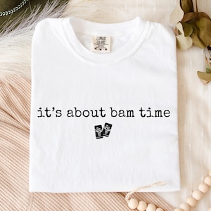 May include: White t-shirt with the phrase "it's about bam time" printed in black, along with two small black graphic images. The shirt is made of a soft material and has a crew neck.