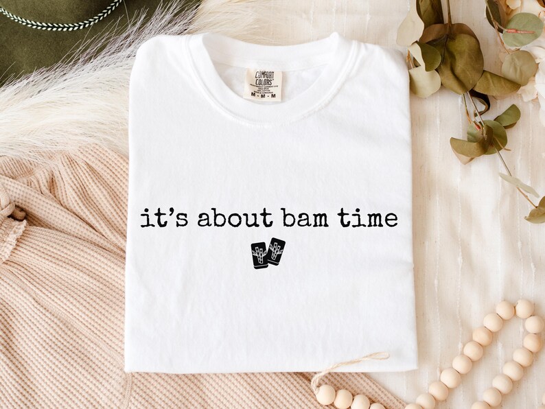 May include: White t-shirt with the phrase "it's about bam time" printed in black, along with two small black graphic images. The shirt is made of a soft material and has a crew neck.