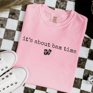 May include: A light pink t-shirt with the phrase "it's about bam time" printed in black. Two small black graphic images are below the text. White canvas sneakers and disco ball ornaments are also visible.