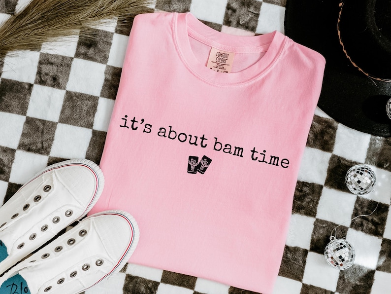 May include: A light pink t-shirt with the phrase "it's about bam time" printed in black. Two small black graphic images are below the text. White canvas sneakers and disco ball ornaments are also visible.