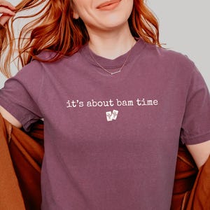 May include: A mauve-colored t-shirt with the phrase "it's about bam time" printed in white. The shirt features a small graphic of two playing cards below the text. The person is wearing a brown jacket.