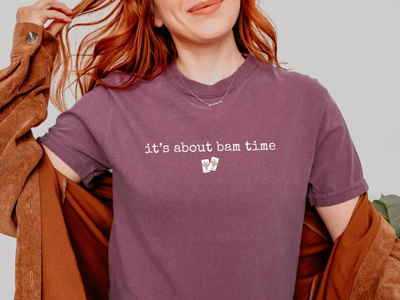 May include: A mauve-colored t-shirt with the phrase "it's about bam time" printed in white. The shirt features a small graphic of two playing cards below the text. The person is wearing a brown jacket.