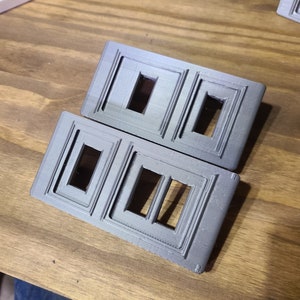 May include: Two gray plastic wall plates with rectangular cutouts for light switches. The plates have a decorative raised border.