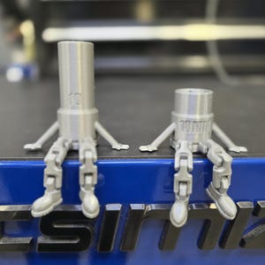 May include: Two silver 3D printed robot figurines sitting on a blue surface with the word "SIMIX" printed in black. The robot on the left has the number "10" printed on its body. The robot on the right has the number "10" and the word "MM" printed on its body.