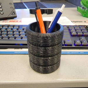 May include: A black tire-shaped pencil holder with five stacked tires. Two pens are in the holder, one orange and one blue.