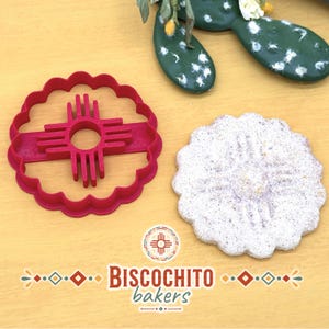 Scalloped flower biscochito cookie cutter with built-in Zia stamp beside finished Zia-imprinted cookie