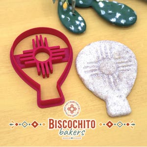 Hot air balloon biscochito cookie cutter with built-in Zia sun stamp beside finished New Mexico style cookie