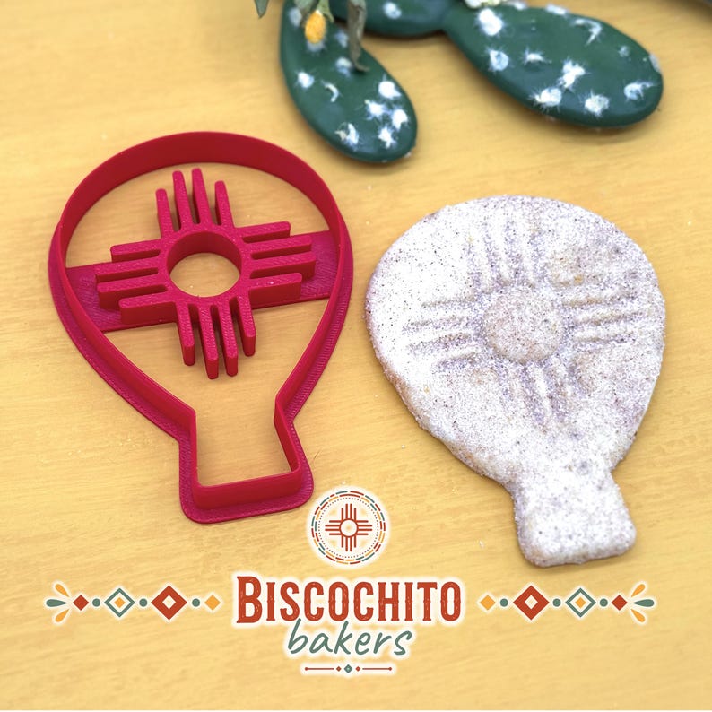 Hot air balloon biscochito cookie cutter with built-in Zia sun stamp beside finished New Mexico style cookie