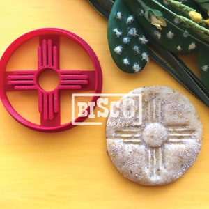 May include: Red plastic cookie cutter in the shape of the Zia symbol, a traditional symbol of the Pueblo people of New Mexico. A cookie with the Zia symbol is also shown.