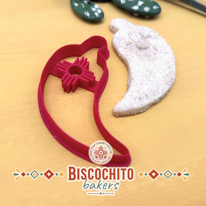 Chile pepper biscochito cookie cutter with built-in Zia sun stamp beside finished New Mexico biscochito