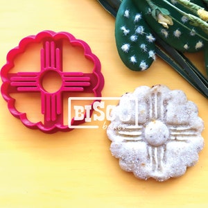 May include: A red plastic cookie cutter shaped like the Zia symbol, a traditional symbol of the Pueblo people of New Mexico. A cookie made with the cutter is shown next to it.