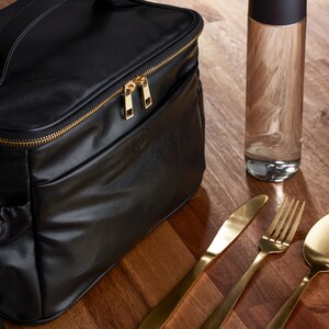 Peut inclure: Sac &agrave; d&eacute;jeuner isotherme noir avec fermeture &eacute;clair dor&eacute;e et logo dor&eacute;. Une bouteille d'eau en verre transparent et des couverts dor&eacute;s sont pos&eacute;s sur une surface en bois. Le sac a une poign&eacute;e et une poche lat&eacute;rale.