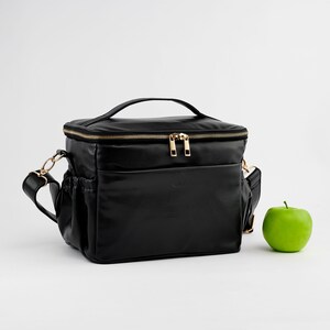 Peut inclure: Sac &agrave; lunch isotherme noir avec fermeture &eacute;clair et quincaillerie dor&eacute;es. Le sac a une poign&eacute;e sup&eacute;rieure et une bandouli&egrave;re amovible. Une pomme verte est pos&eacute;e &agrave; c&ocirc;t&eacute; du sac. Le sac a une poche avant et des poches lat&eacute;rales.