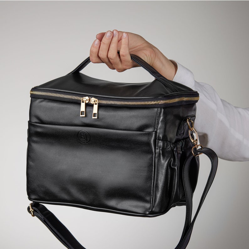 Peut inclure: Sac noir rectangulaire avec une poign&eacute;e sup&eacute;rieure et une bandouli&egrave;re amovible. Le sac est dot&eacute; d'une fermeture &eacute;clair et d'une quincaillerie dor&eacute;es. Un petit logo circulaire est visible sur le devant. Le sac est tenu par une personne.