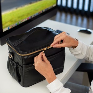 Peut inclure: Sac de maquillage en cuir noir avec une fermeture &eacute;clair et une poign&eacute;e dor&eacute;es. Le sac a une forme rectangulaire avec une poche lat&eacute;rale et une bandouli&egrave;re. La fermeture &eacute;clair est ouverte.