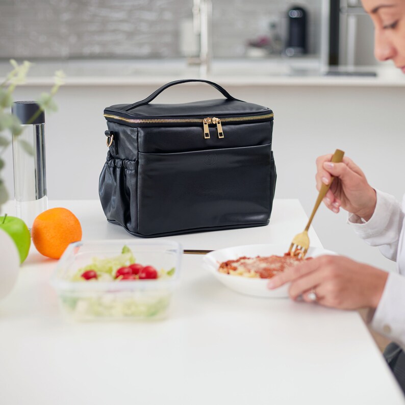 Peut inclure: Sac &agrave; d&eacute;jeuner noir avec une fermeture &eacute;clair et une poign&eacute;e dor&eacute;es. Le sac a une bandouli&egrave;re et des poches lat&eacute;rales. Une orange, une pomme verte et un r&eacute;cipient de salade sont sur la table. Une personne mange des p&acirc;tes avec une fourchette dor&eacute;e.