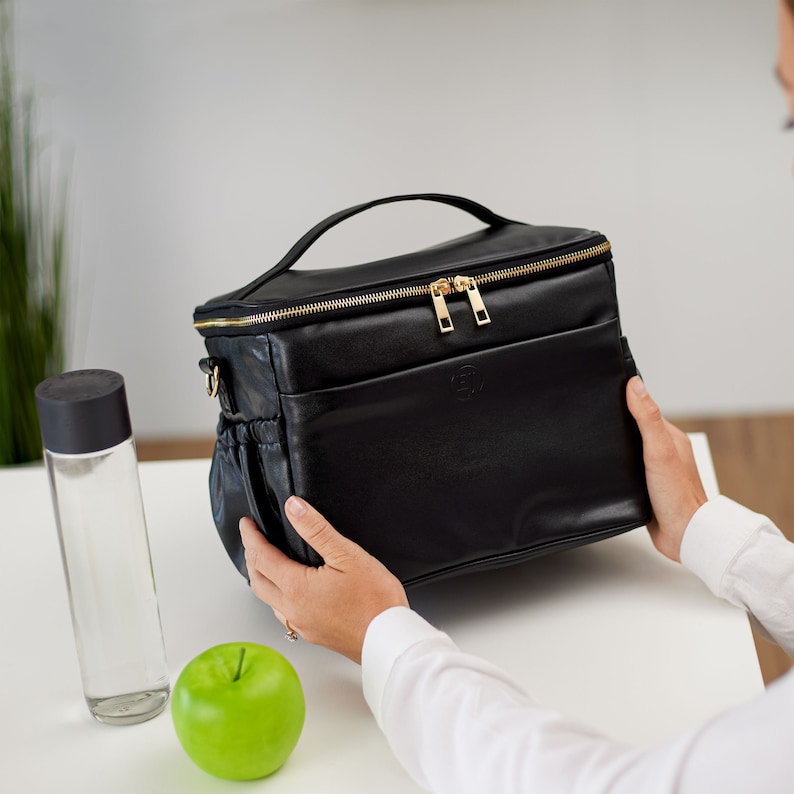 Peut inclure: Sac &agrave; d&eacute;jeuner noir avec une fermeture &eacute;clair et une poign&eacute;e dor&eacute;es. Le sac a une poche avant et une poche lat&eacute;rale. Une bouteille d'eau transparente avec un couvercle noir et une pomme verte sont &agrave; c&ocirc;t&eacute; du sac.