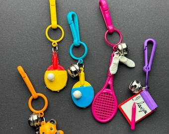 May include: Five colorful keychains with different charms. The charms include a tennis racket, a ping pong paddle, a teddy bear, a notepad with a pencil, and a pair of sneakers. Each keychain has a small bell attached.