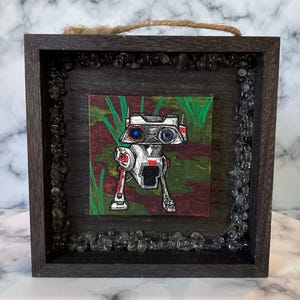 May include: Square wooden framed artwork featuring a robot illustration. The robot is white, red, and black, set against a green and red background. The frame is dark brown, with clear stones around the inner edge and a jute rope for hanging.