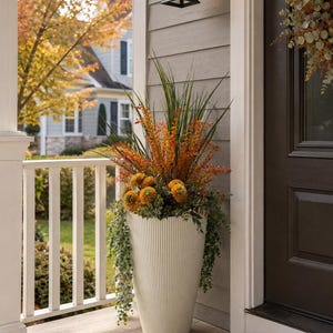 May include: A tall, off-white, ribbed cylindrical planter filled with an arrangement of artificial greenery, including long green grass blades, orange floral sprays, round orange blossoms, and trailing eucalyptus-style foliage.