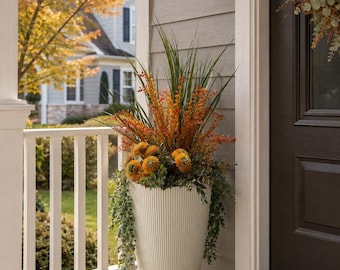 Peut inclure: Un grand pot cylindrique cannelé blanc cassé rempli d'un arrangement de feuillage artificiel d'automne, comprenant des grappes de fleurs orange, de hautes herbes vertes et des feuilles d'eucalyptus tombantes.