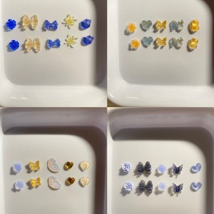 Könnte beinhalten: Sortiment an bunten Harz-Anhängern in verschiedenen Formen, darunter Schmetterlinge, Herzen, Sterne und Blumen. Die Anhänger sind in vier separaten Behältern auf weißem Hintergrund angeordnet.