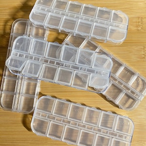 Puede incluir: Cuatro contenedores de plástico transparente con compartimentos individuales para guardar objetos pequeños. Cada contenedor tiene una tapa con bisagras y está etiquetado con números.