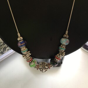 May include: A silver chain necklace with colourful glass beads and a silver charm. The beads are various shades of blue, green, red, and white. The charm is a silver flower with intricate details.