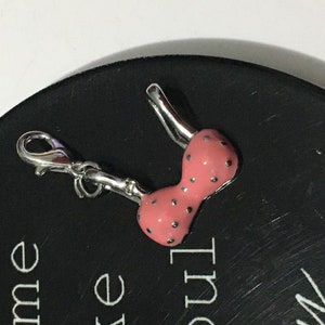 Könnte beinhalten: Ein silberner Anhänger in Form eines rosa BHs mit weißen Punkten. Der Anhänger hat einen silbernen Verschluss.