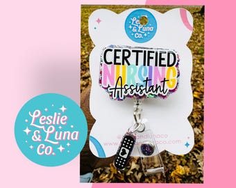 May include: A colourful glitter badge reel with the text "CERTIFIED NURSING ASSISTANT". The reel is attached to a silver chain with a black and white bandage charm and a purple glitter charm.