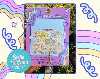 May include: A badge reel with a colourful design. The badge reel has the text "SOMEONE'S WEIRD COWORKER" in a playful font. The background is a light blue with a star and heart shape. The badge reel is attached to a card with the Leslie and Luna Co. logo.