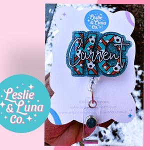 May include: A blue and white glitter badge reel with the text "Current" and soccer ball designs. The reel is attached to a clear plastic retractable cord with a silver clip. The background is white with the text "Leslie & Luna Co."