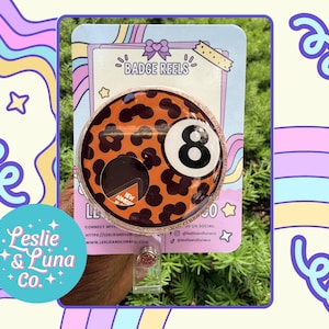 May include: A circular badge reel featuring an orange background with a black leopard print pattern and a white number eight billiard ball graphic. The badge reel is mounted on a light blue display card with the text BADGE REELS at the top.