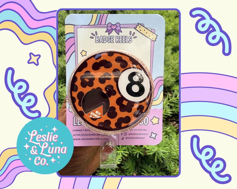 May include: A circular badge reel featuring an orange background with a black leopard print pattern and a white number eight billiard ball graphic. The badge reel is mounted on a light blue display card with the text BADGE REELS at the top.