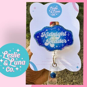 May include: A badge reel with a blue and glittery cloud-shaped design, featuring the words "Midnight Reader" in a retro font. The badge reel is attached to a white card with the "Leslie & Luna Co." logo.