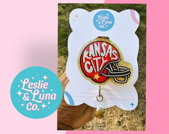 May include: A badge reel featuring a red and white football helmet graphic with the text Kansas City in bold lettering. The helmet is set against a gold glitter background on a white display card with the Leslie & Luna Co. logo at the top.