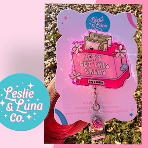 May include: Pink and white badge holder with a pink and white toaster with the text "Let's Get This Bread" and the text "Not a Fresh" on the bottom. The badge holder has a pink glitter design and a pink and white star design.