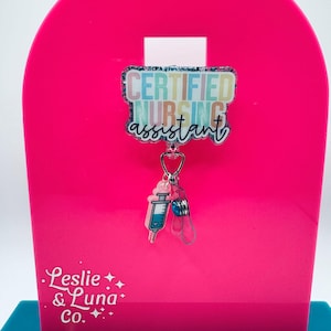 May include: A badge reel featuring a rectangular acrylic topper with a glittery finish and the text CERTIFIED NURSING assistant in colourful block and script lettering. A small pink syringe charm and a blue bead dangle from the central clip.