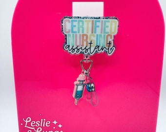 May include: A badge reel featuring a rectangular acrylic topper with a glittery finish and the text CERTIFIED NURSING assistant in colorful block and script lettering. A small pink syringe charm and a blue bead dangle from the central clip.