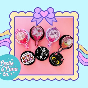May include: A collection of decorative hand mirrors and round pins. The mirrors feature pink bows and various designs, including a cat, a butterfly, and the text "GIVE ME MORE BOOKS PLS THX". The background is light blue and pink.