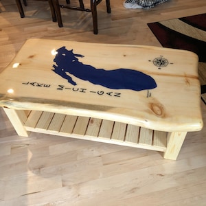 May include: A wooden coffee table with a live edge top and a lower shelf. The top is stained with a clear epoxy resin and features a blue outline of Lake Michigan with the text "LAKE MICHIGAN" underneath. A compass rose is also visible on the top.