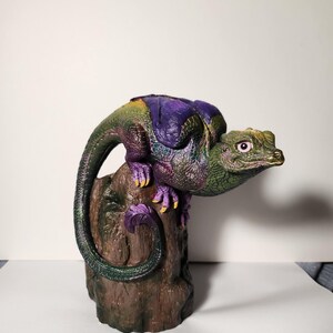 May include: A small, green, purple, and yellow dragon figurine perched on a brown, wooden stump. The dragon has a long tail that wraps around the stump.