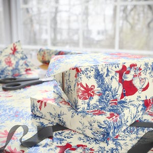 May include: Two rectangular gift boxes wrapped in cream-colored paper featuring a repeating toile-style pattern of Santa Claus, red bows, and blue pine branches. A pair of black scissors and a black ribbon are placed on the patterned wrapping paper.