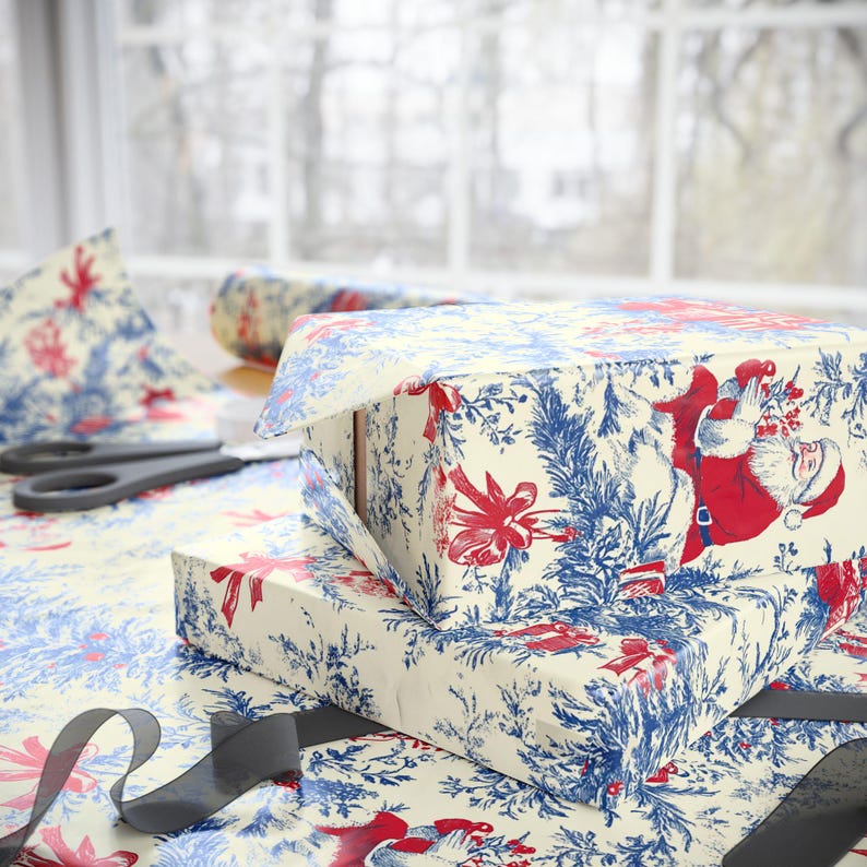 May include: Two rectangular gift boxes wrapped in cream-colored paper featuring a repeating toile-style pattern of Santa Claus, red bows, and blue pine branches. A pair of black scissors and a black ribbon are placed on the patterned wrapping paper.