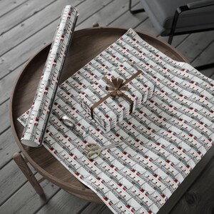 K&ouml;nnte beinhalten: Eine Rolle wei&szlig;es Geschenkpapier und ein Geschenk mit braunem Band, beide mit einem sich wiederholenden Muster aus grauen und roten Kreuzfahrtschiffen. Goldene Schere und Klebeband liegen auf dem Holztisch.